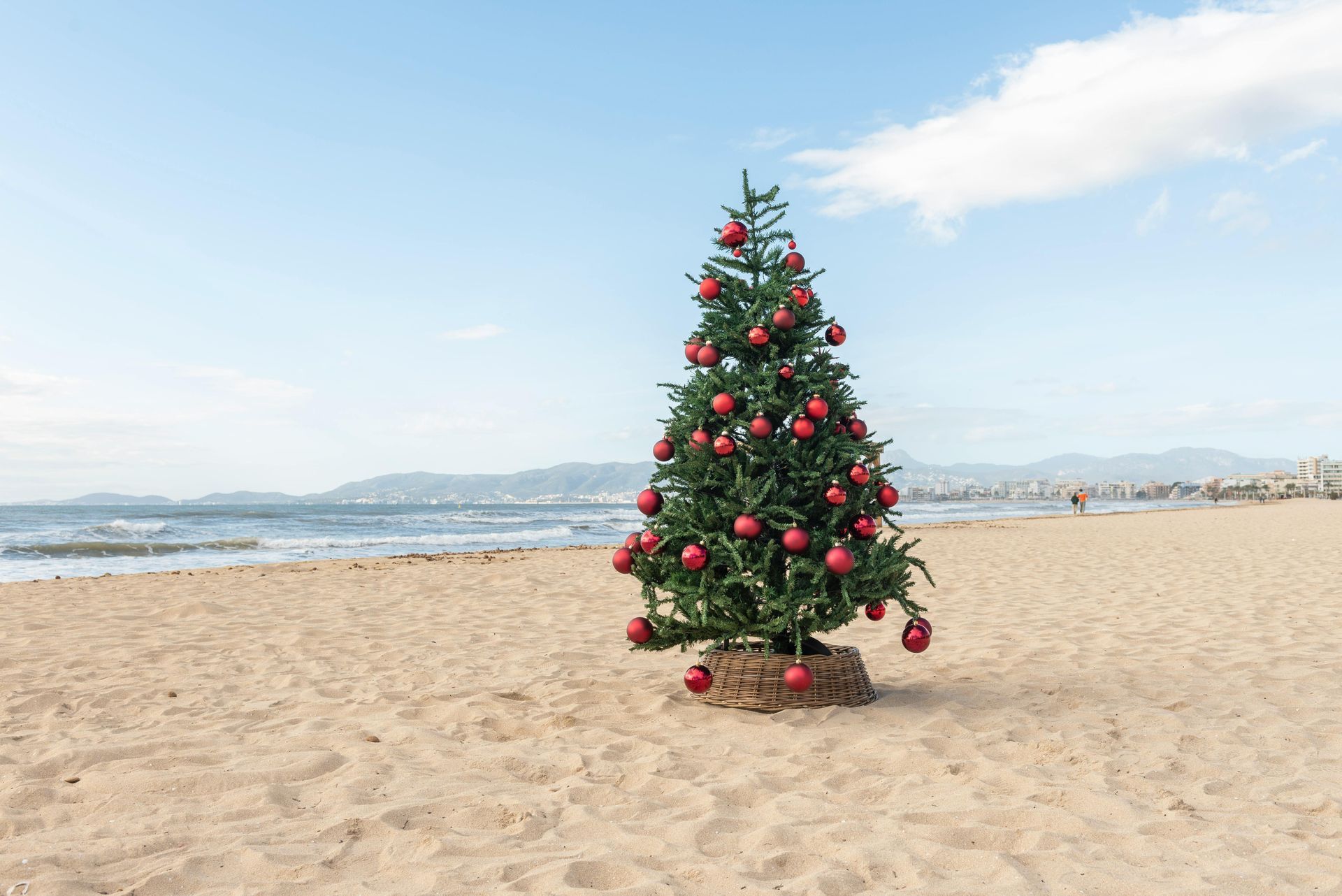 Christmas tree decorated with red ornaments on a sandy beach, ocean in background.