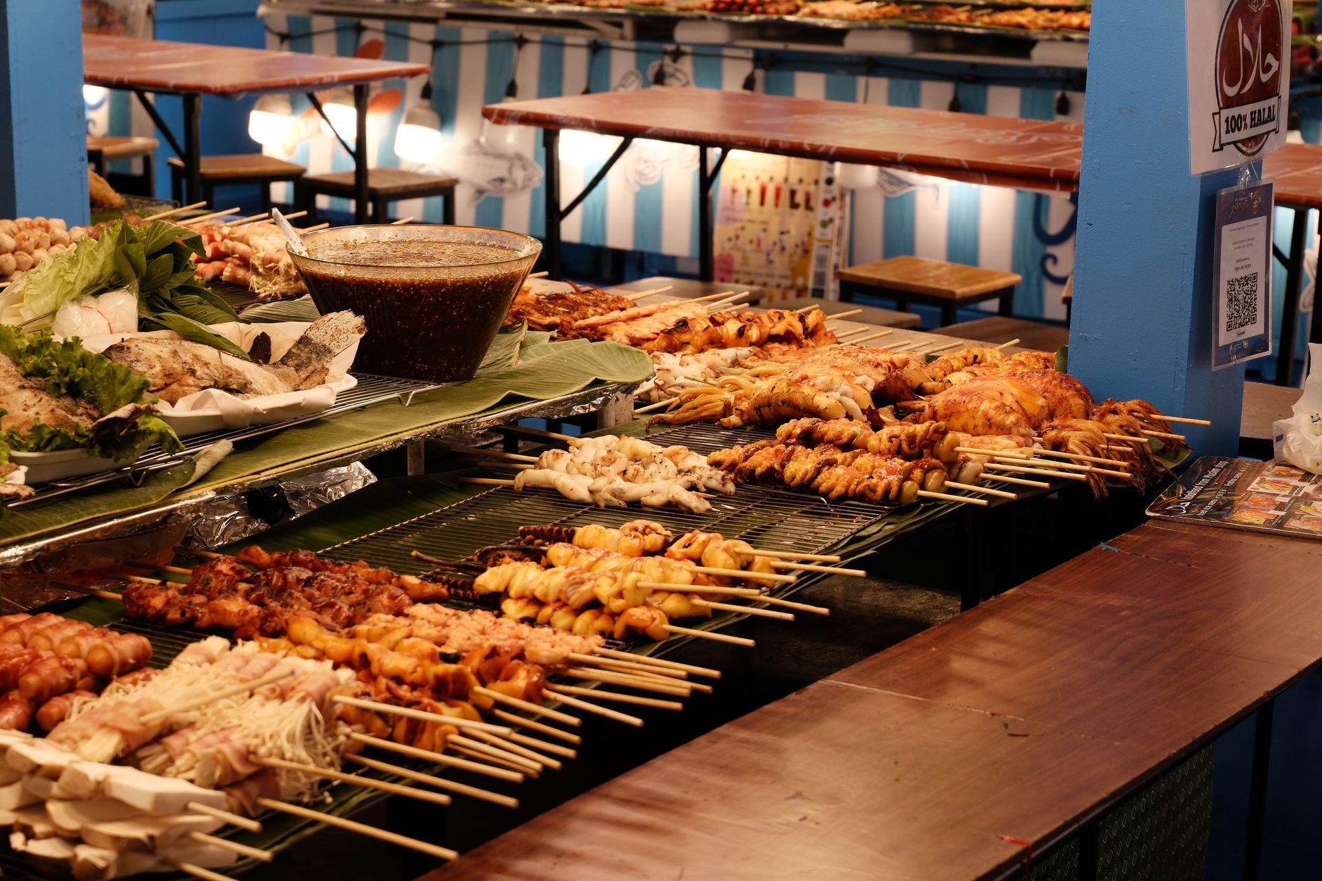 Grilled skewers and seafood arranged on market-style buffet trays with tongs on a wooden table.