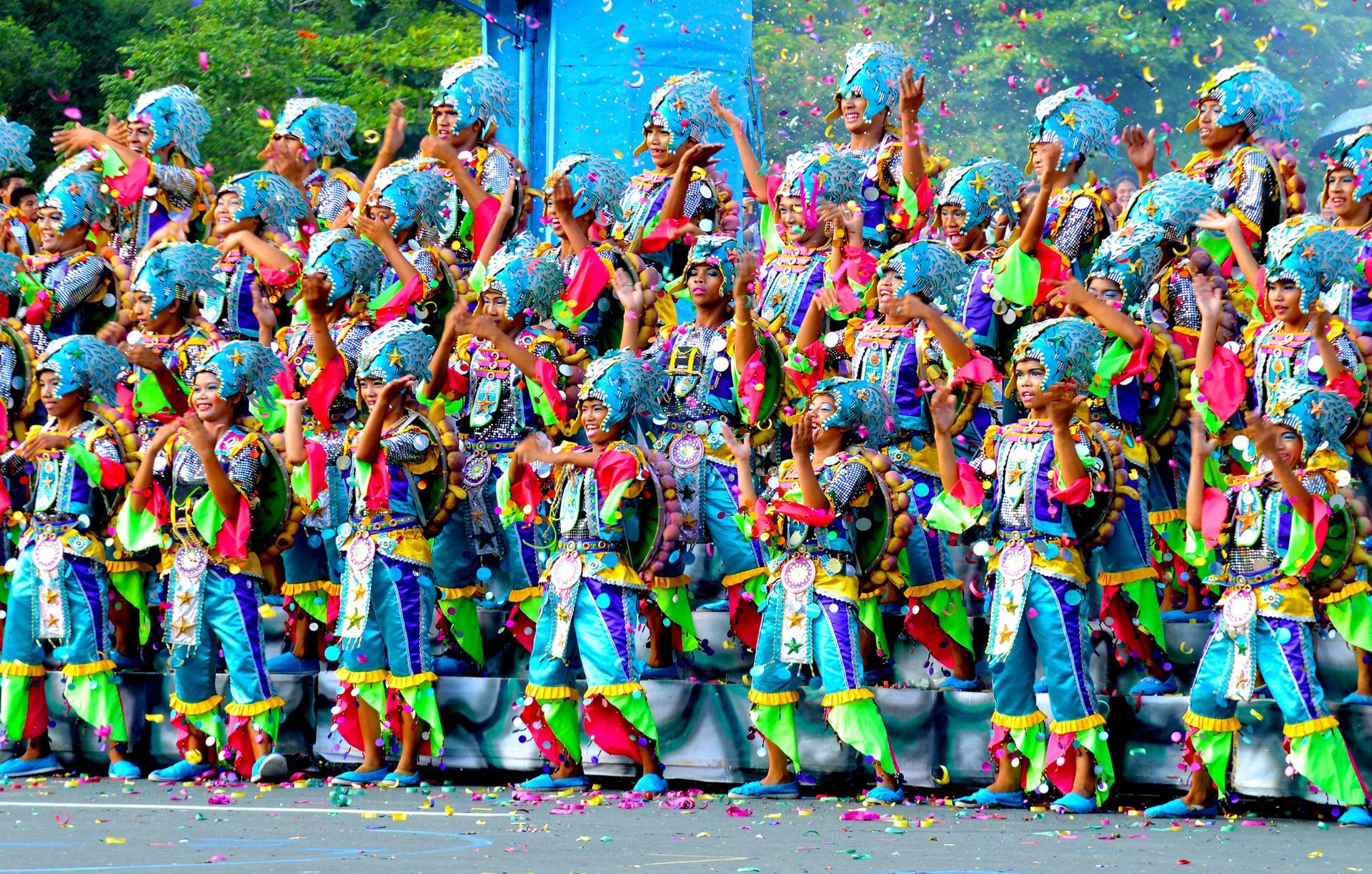 Colorfully dressed performers in a street parade dancing and waving flags before a blue backdrop