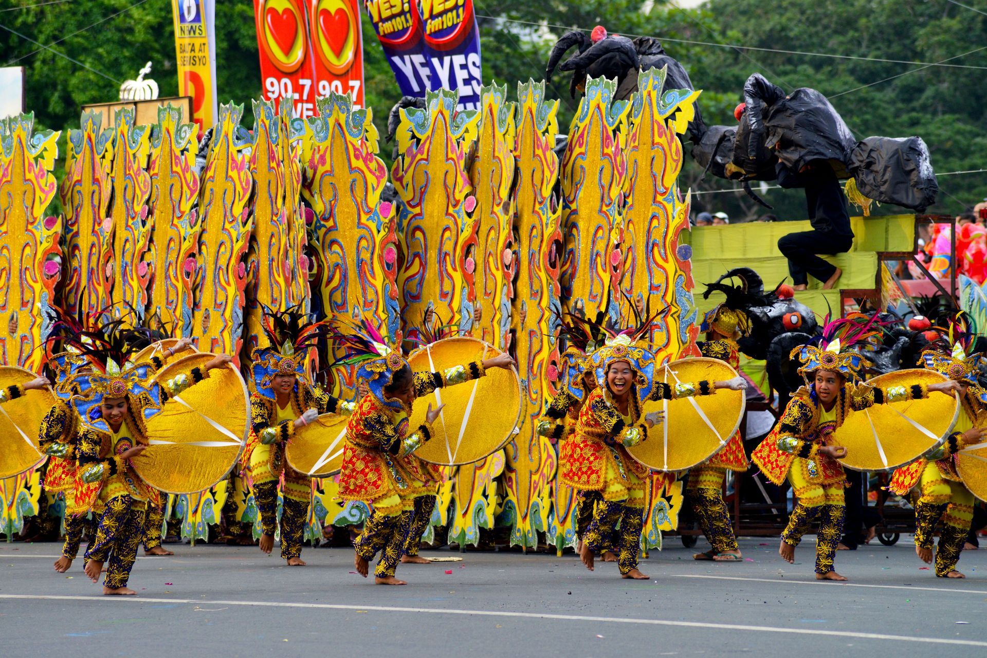 Colorful parade of dancers in gold costumes with yellow props and tall banners marching on a street