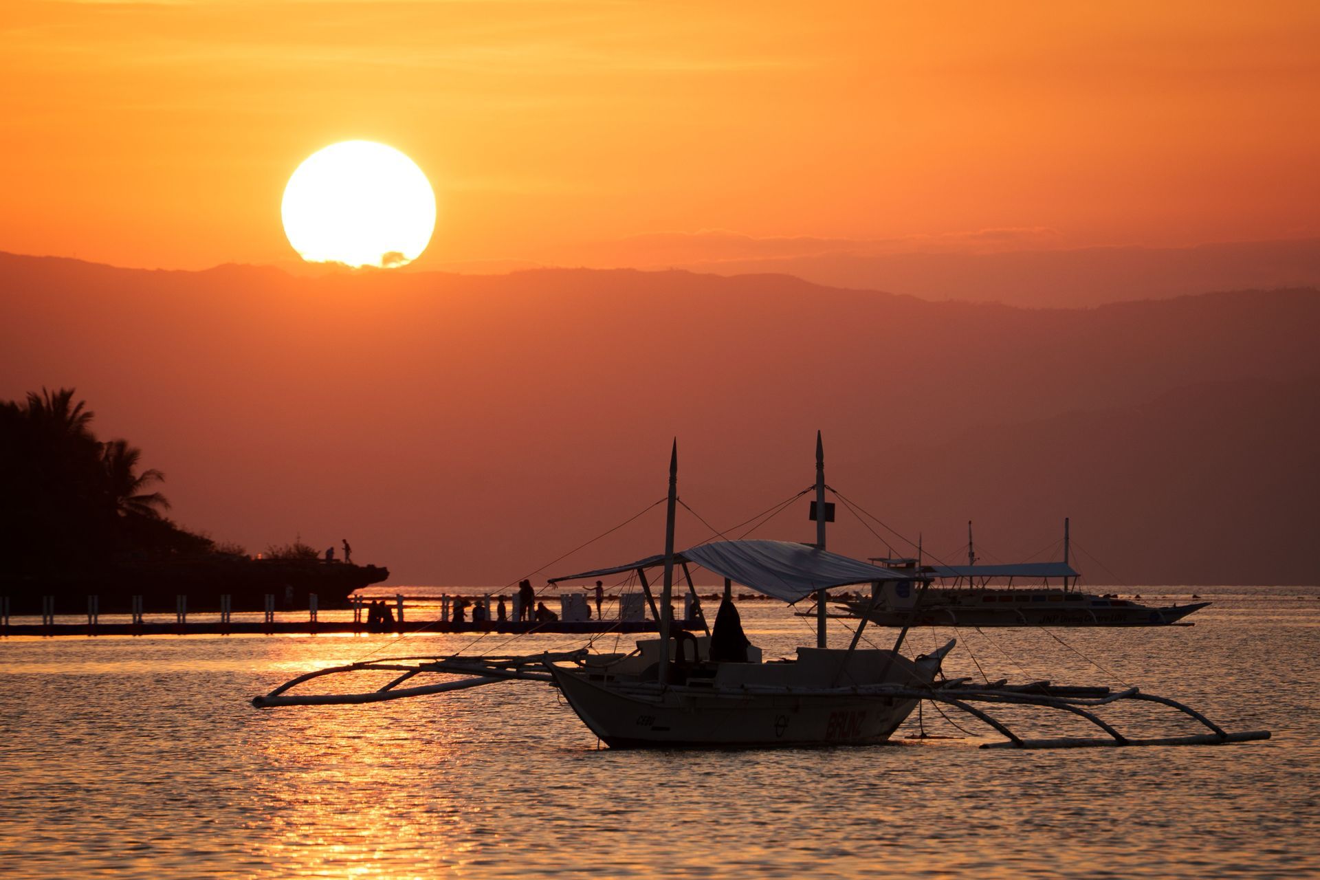 Sunset over calm water with a silhouetted outrigger boat and pier