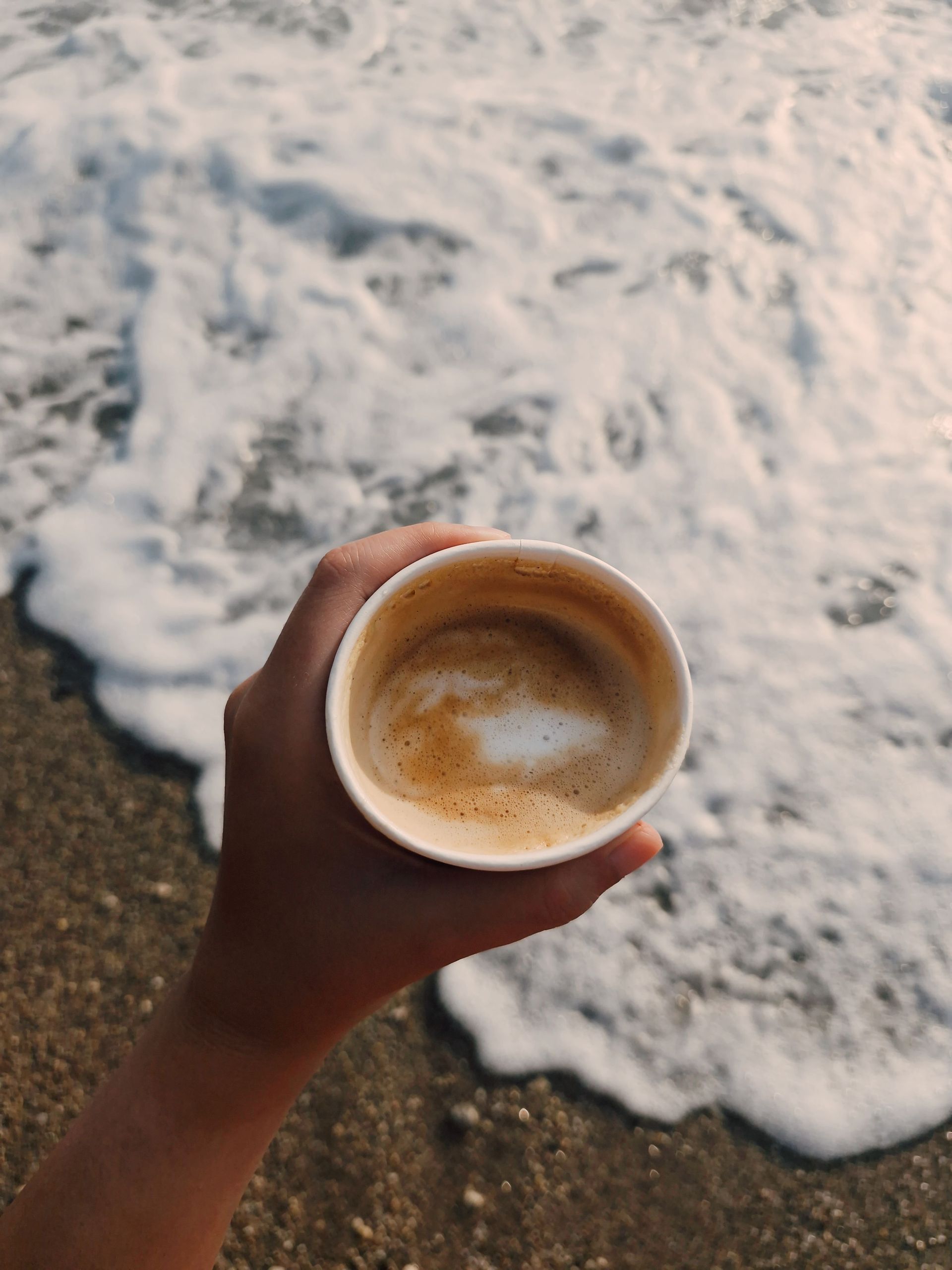 Hand holding a cup of coffee beside foamy ocean waves on a sandy beach