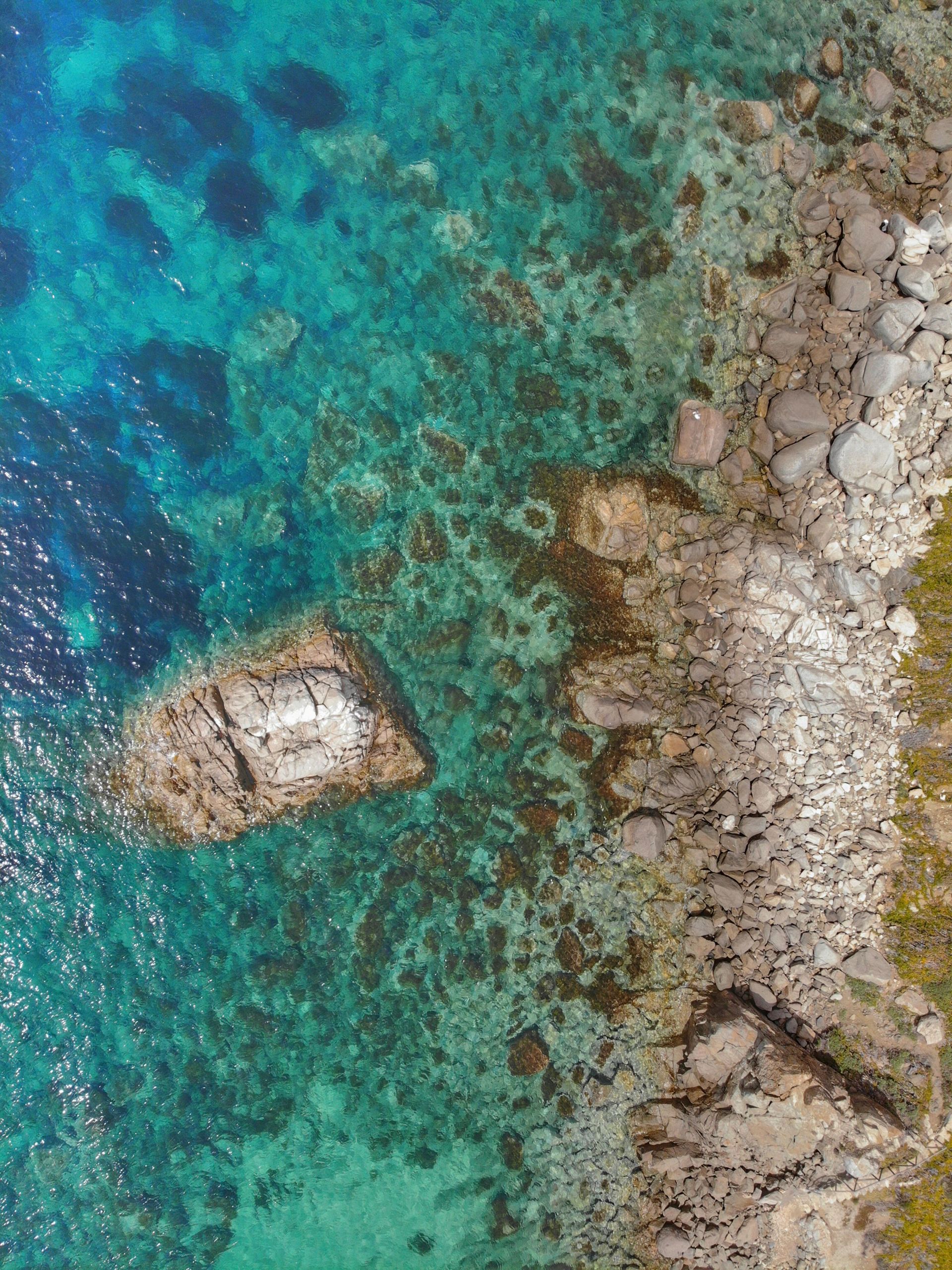 Aerial view of turquoise water beside a rocky shoreline with a large rock in the shallows