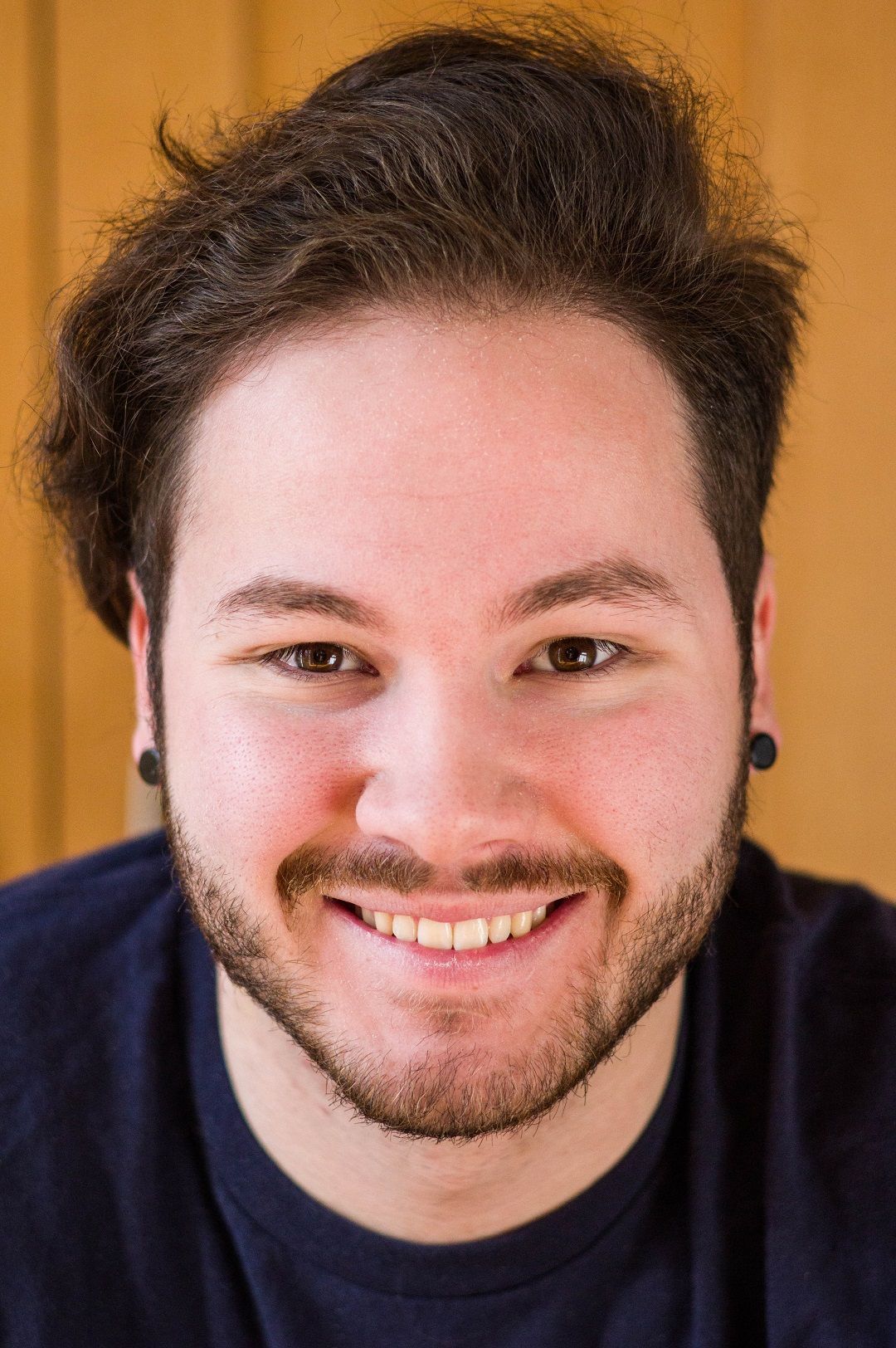 Man with dark hair and beard smiles, wearing a blue shirt, with gauges in his ears.