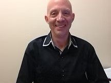 Bald man with a friendly smile wearing a black shirt with white trim, indoors.