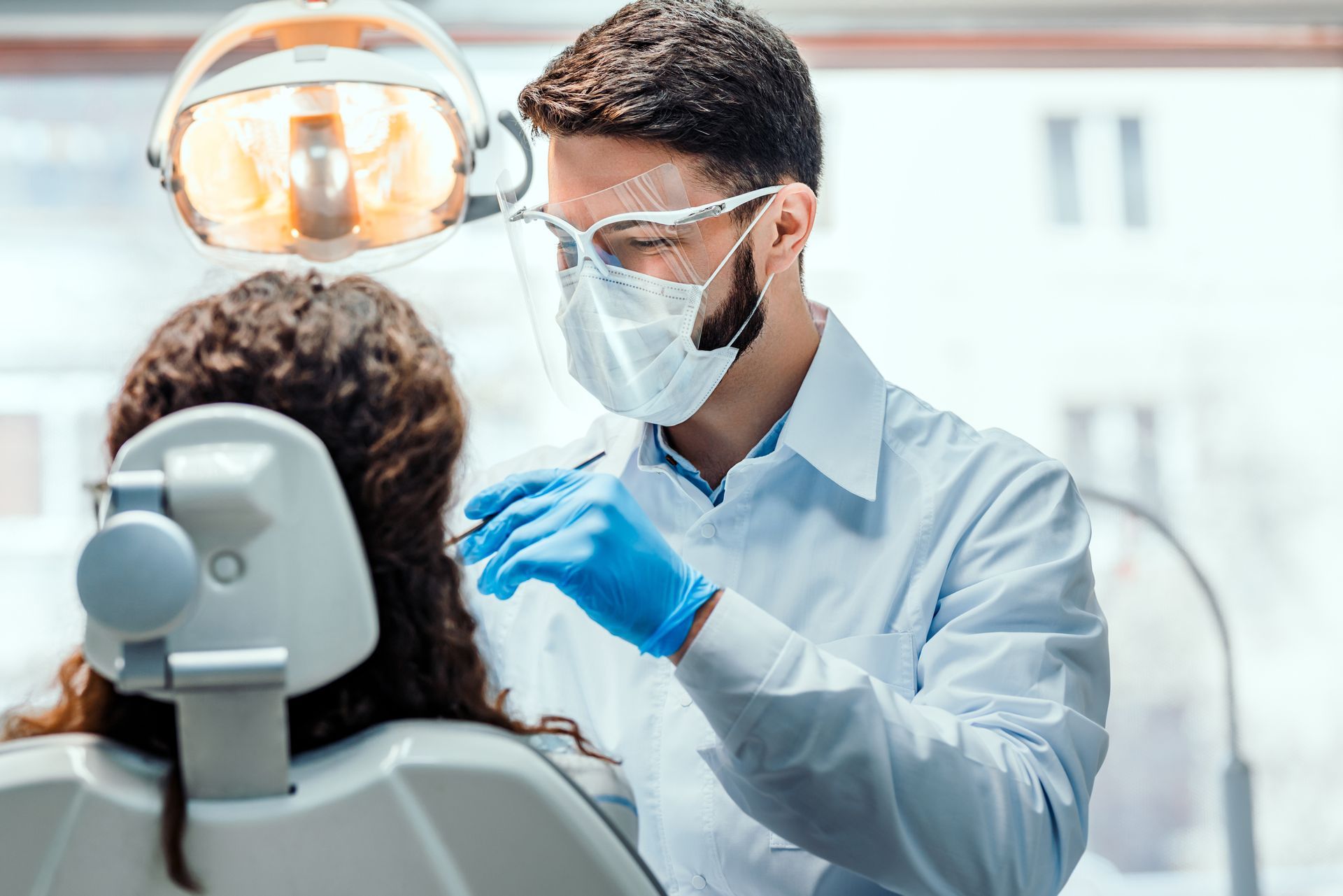 Dentist wearing mask and gloves examines a patient in a dental chair under bright clinic lights