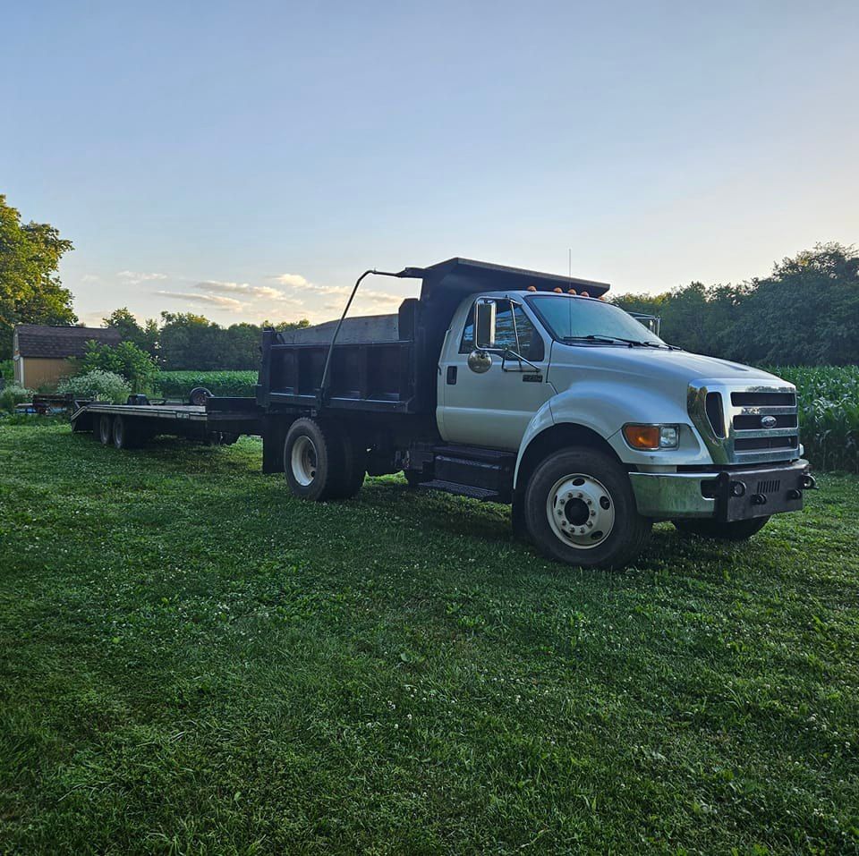 A silver dump truck with a flatbed trailer attached, parked on a grassy field under a clear sky.