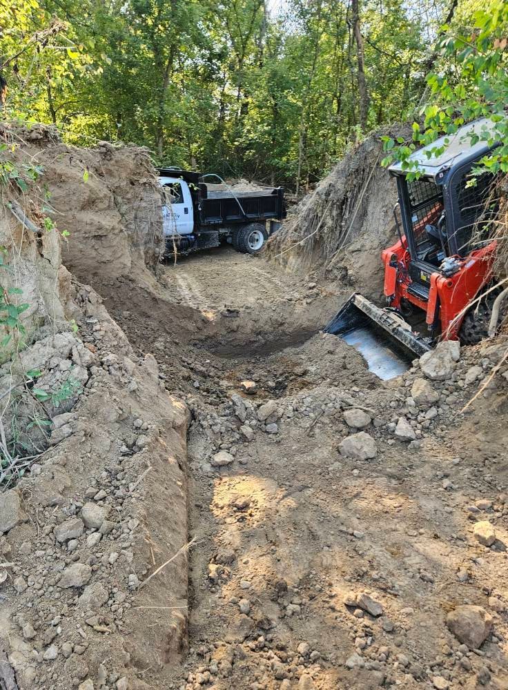 A white dump truck and a red skid steer loader parked in a deep, excavated dirt trench surrounded by trees.