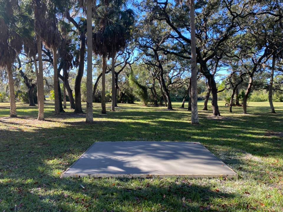 Concrete pad in a sunny park, surrounded by trees and grass, likely for a disc golf course.