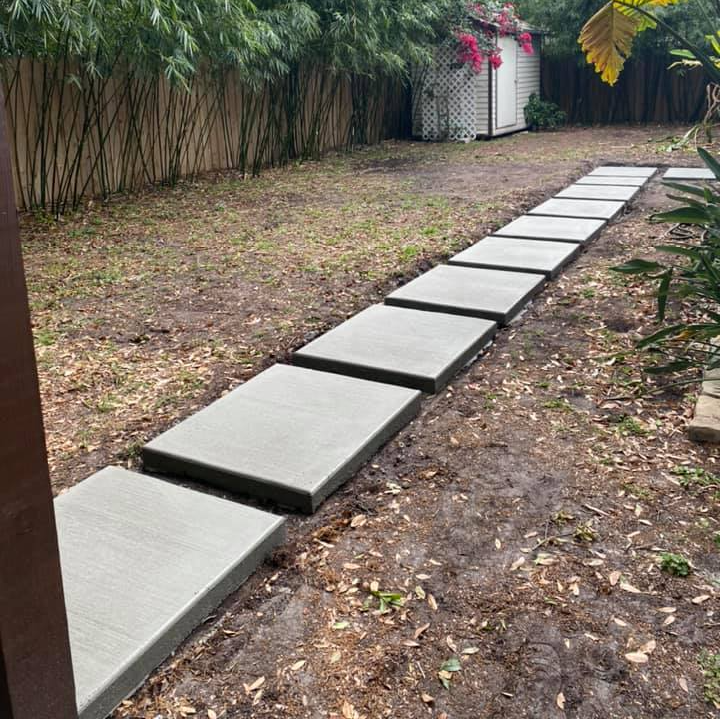 Concrete stepping stones in a backyard lead to a white shed with pink flowers, edged by bamboo.