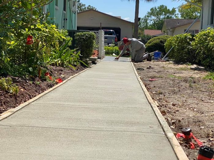 Man kneels, smoothing wet concrete sidewalk in front yard. Gray cement, wood borders, green bushes. Sunny day.