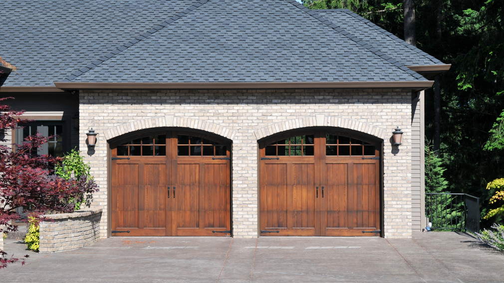 Two wooden garage doors on a stone house with a dark shingled roof and driveway