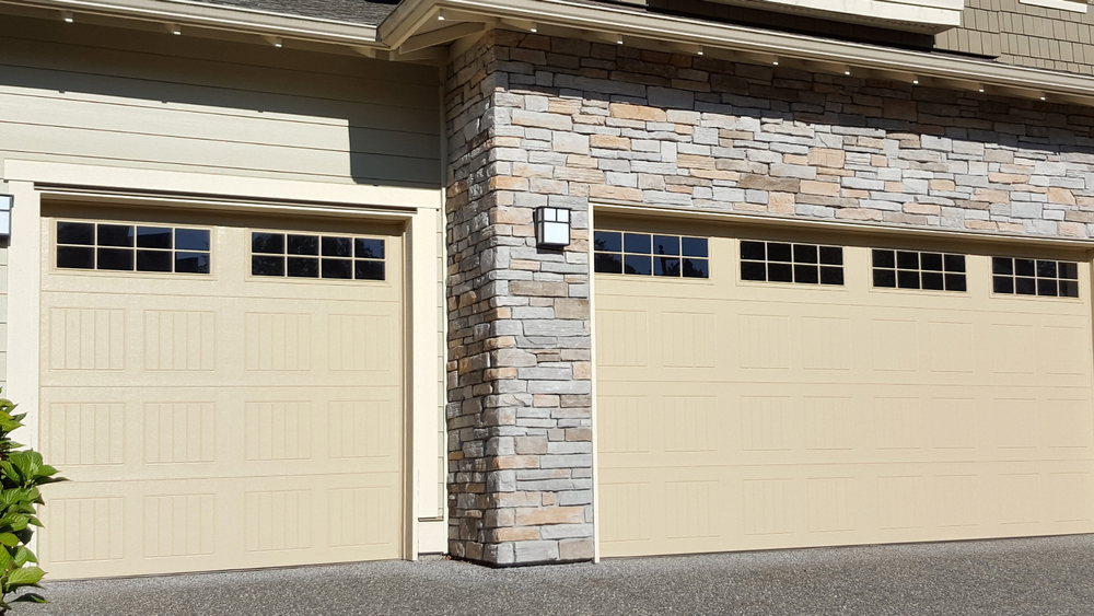 Two beige garage doors with small windows on a stone and siding house exterior
