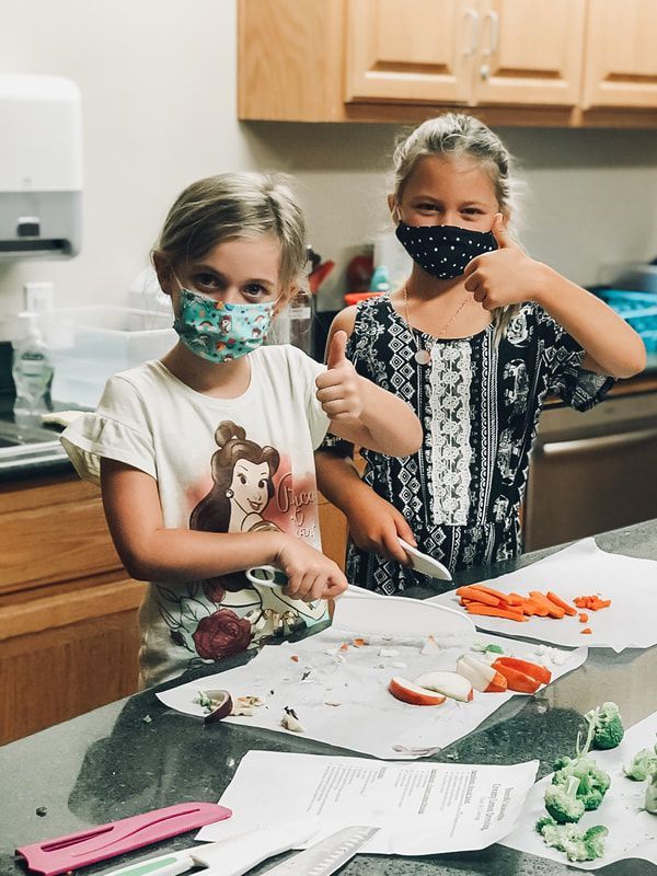 Two young girls wearing face masks are cutting vegetables and giving a thumbs up.