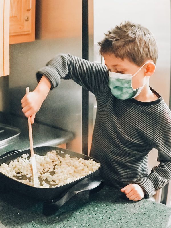 A young boy wearing a mask is stirring food in a pan.