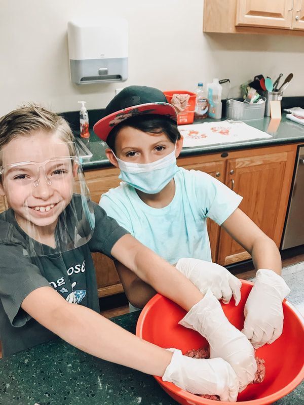 Two young boys wearing face masks and gloves are mixing something in a red bowl.