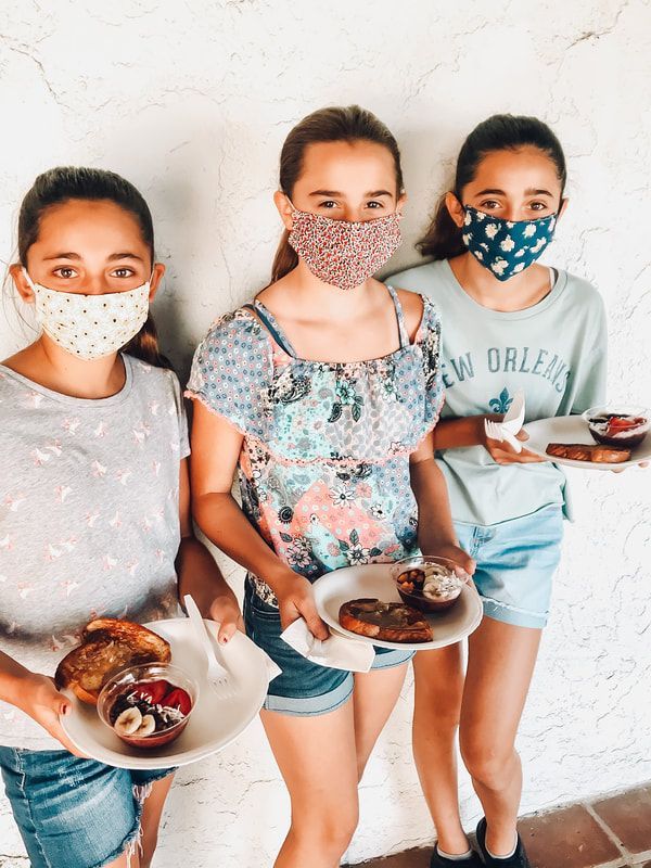 Three girls wearing face masks are holding plates of food.