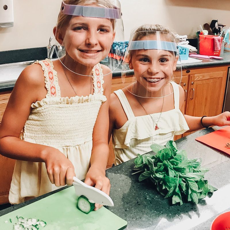 Two young girls wearing face shields are preparing food in a kitchen