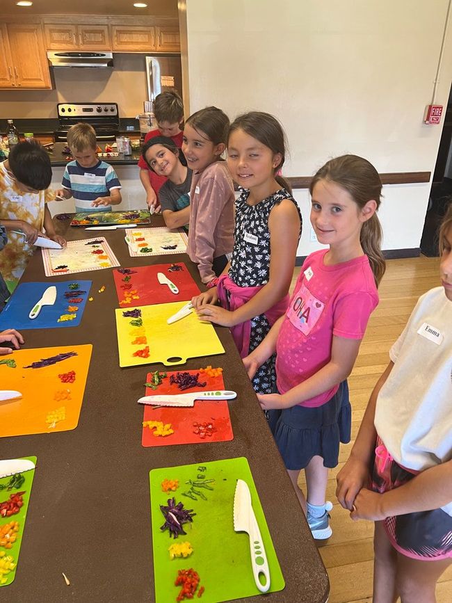 A group of children are standing around a table with knives and cutting mats.