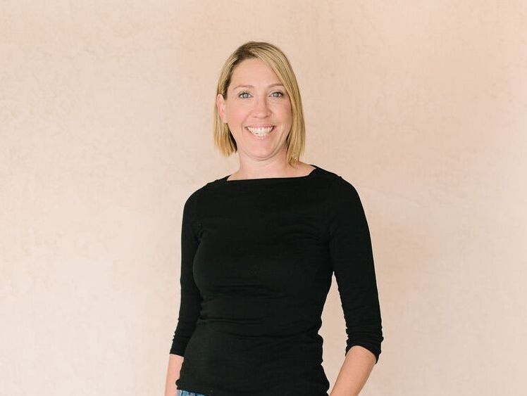 A woman in a black shirt is smiling and standing with her hands in her pockets.