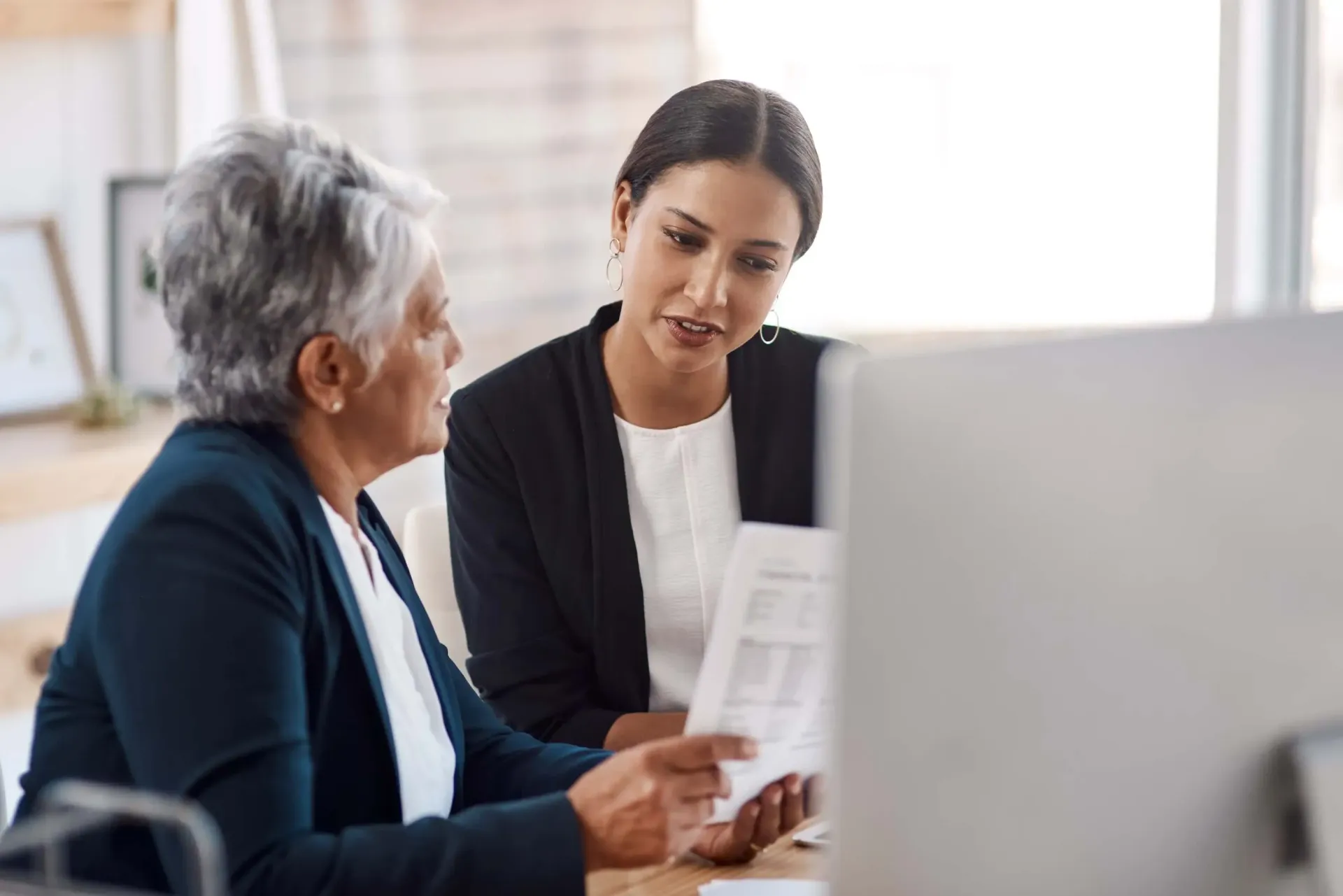 Two women in an office discussing paperwork at a computer workstation