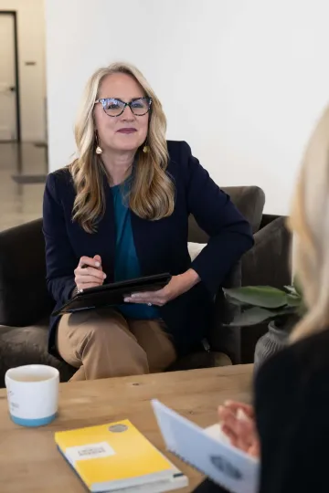 Woman with glasses holding a tablet in a meeting room, speaking to someone across a table.