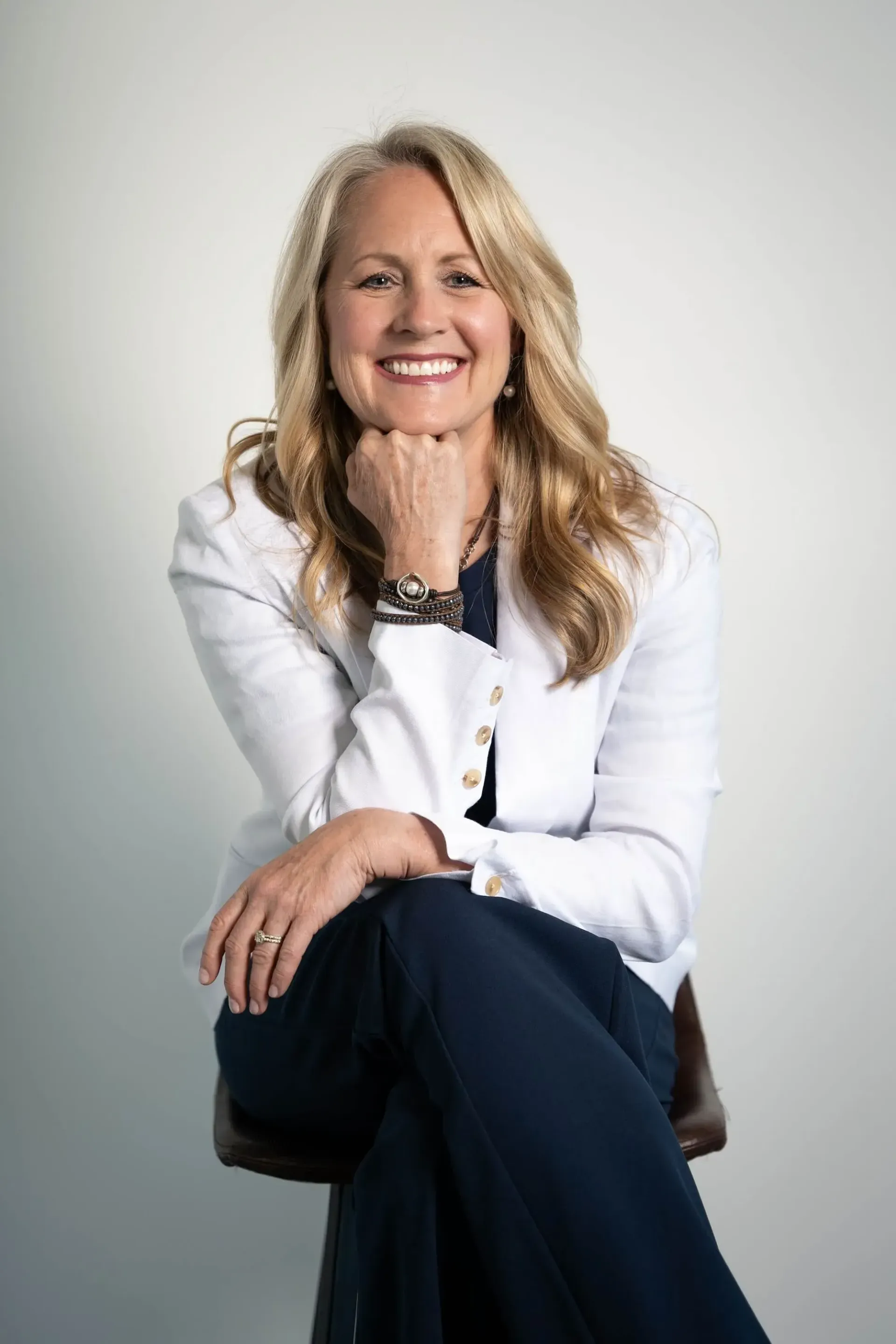 Smiling woman in a white blazer seated against a light gray background, resting her chin on her hand
