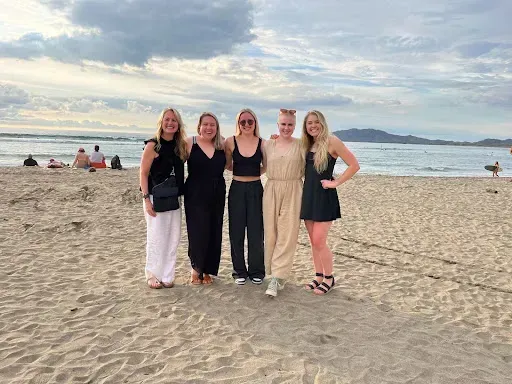 Five friends posing on a sandy beach at sunset, with the ocean and mountains in the background.