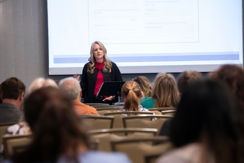Speaker presenting to seated audience in a conference room with a projected slide behind her