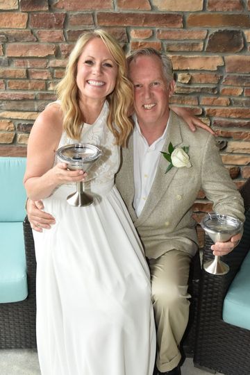 Smiling couple in wedding attire seated on a couch, holding champagne glasses against a brick wall.