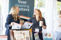 Three people at a booth chatting under a “Your meaning matters” banner, holding pamphlets and a coffee cup