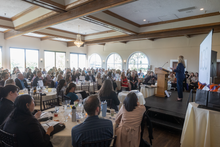 Conference banquet with seated audience and a speaker on a stage in a bright hall