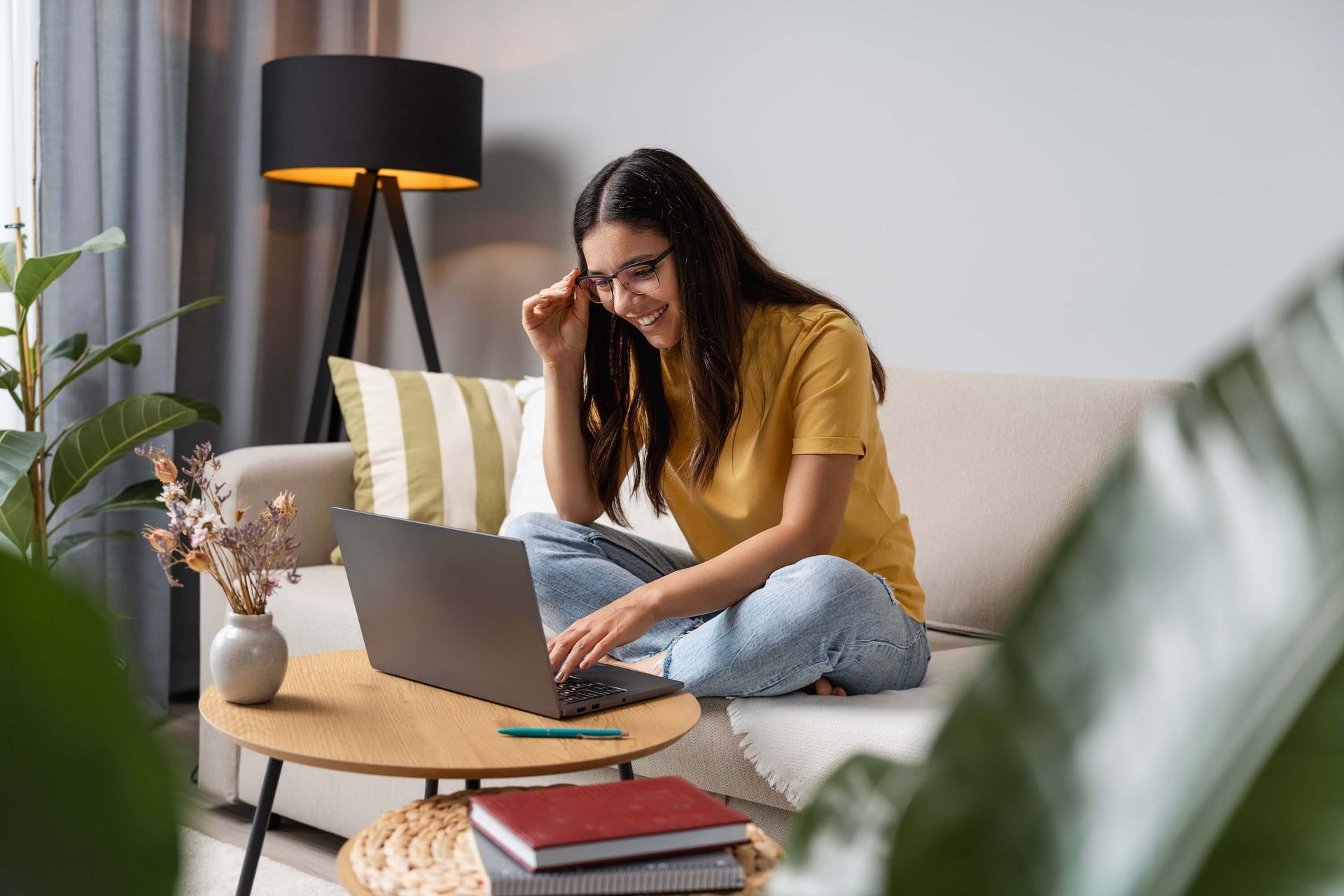 Woman working on a laptop on a sofa in a bright living room with a lamp and plants.