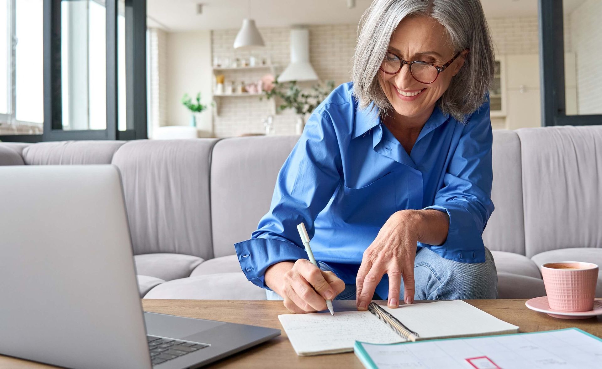 Woman in blue shirt smiling at a laptop on a living room sofa, writing in a notebook.