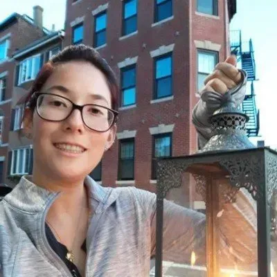 Person smiling beside a lit lantern in front of a brick building at dusk