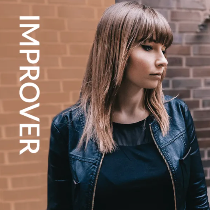 Woman in a black jacket against a brick wall with the word “IMPROVER” beside her