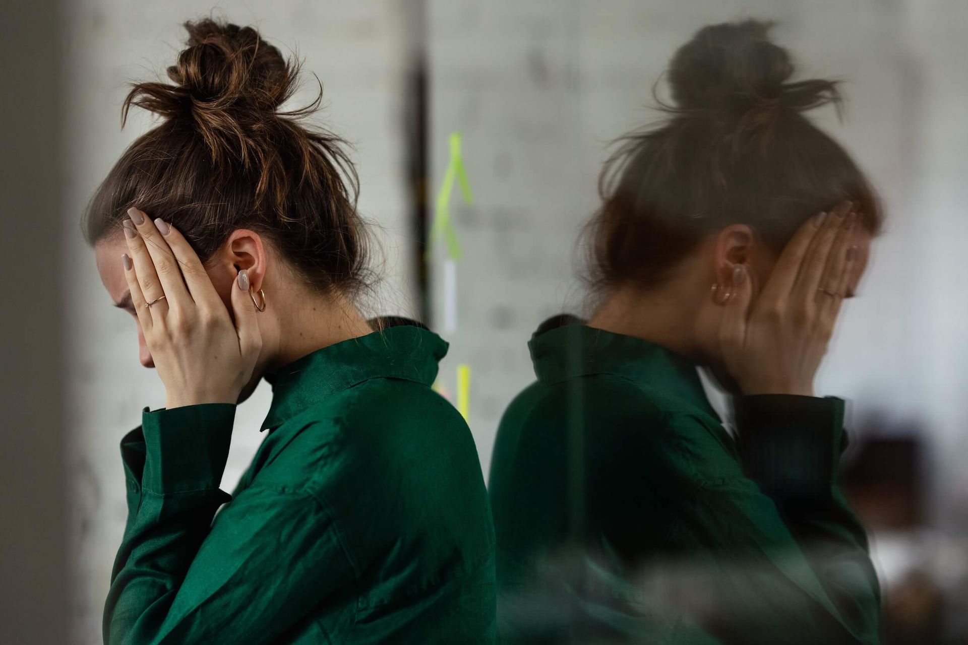Person in a green jacket covers their face, reflected in a mirror with a blurred double image.
