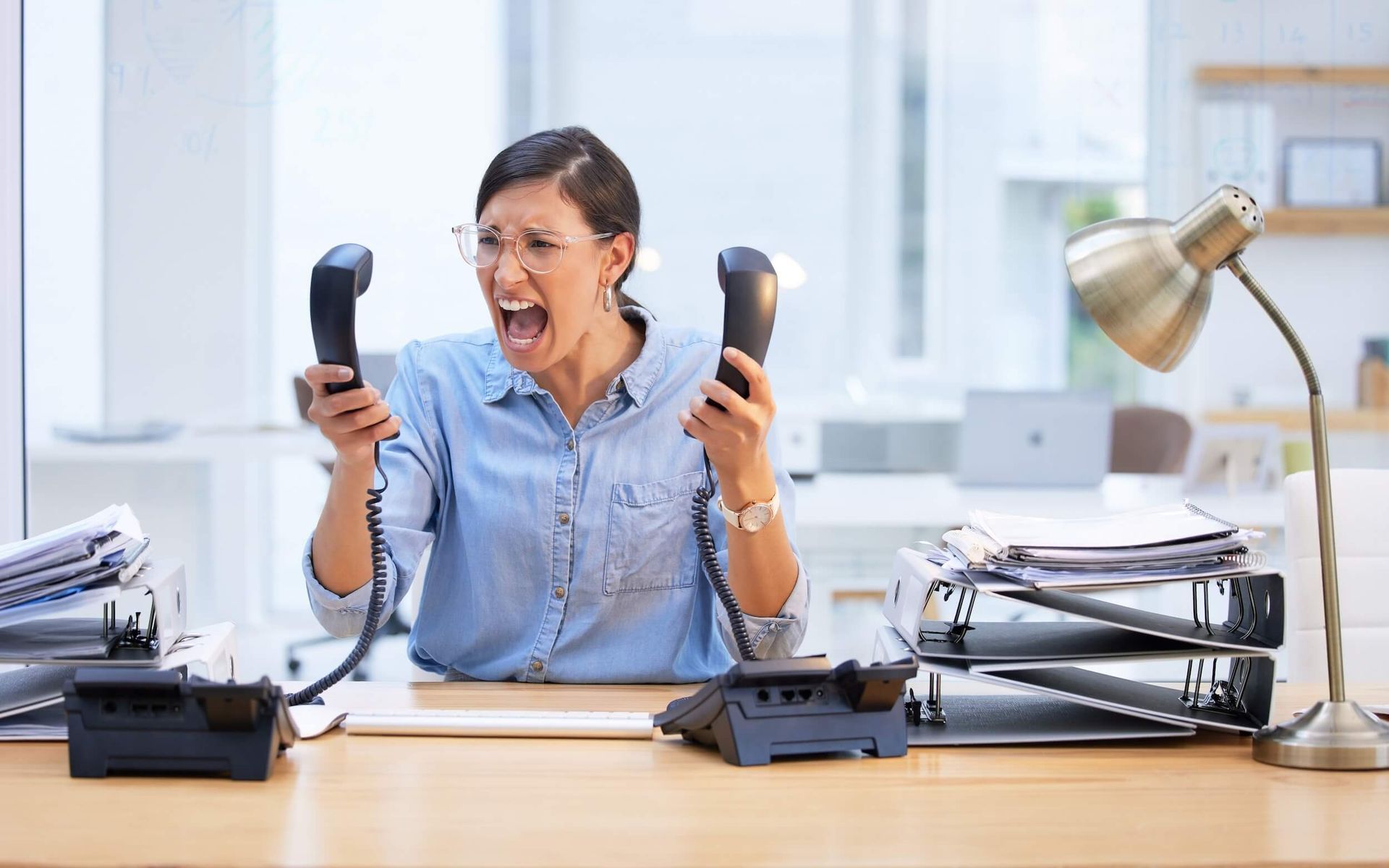 Frustrated office worker shouting while holding two phones at a cluttered desk
