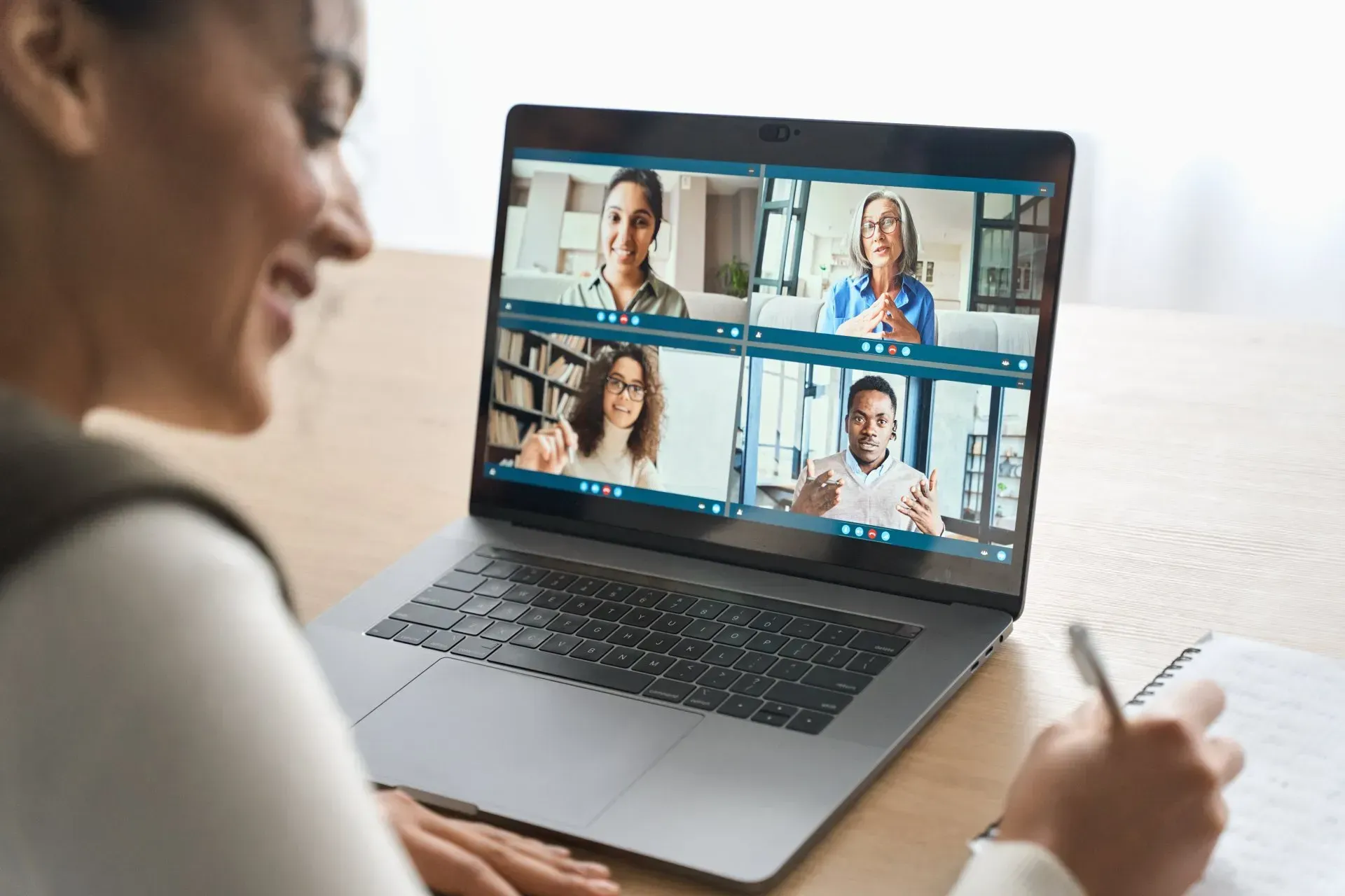 Person on a laptop video call with four participants in a grid view, taking notes at a desk.