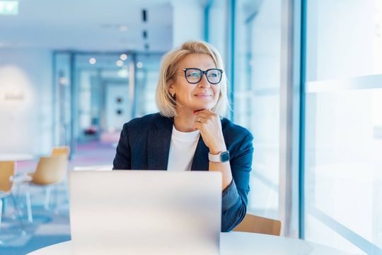 Woman with glasses working on a laptop in a bright office, looking out the window задумленно