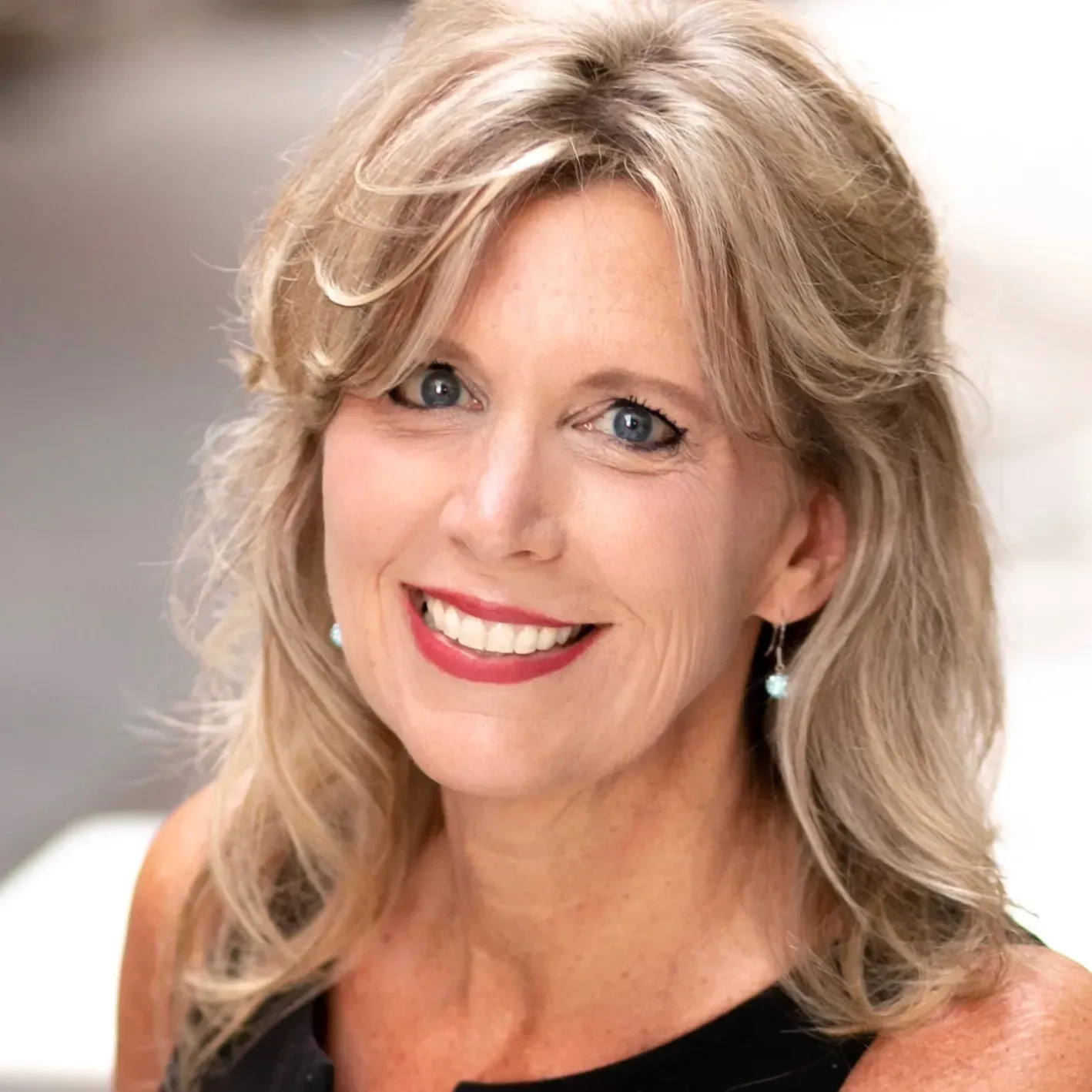 Smiling woman with blonde hair and red lipstick against a soft gray background