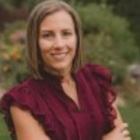 Woman in a burgundy blouse smiling outdoors with blurred greenery in the background