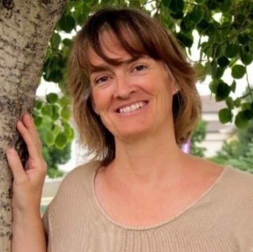 Smiling woman in a beige top standing by a tree with green leaves
