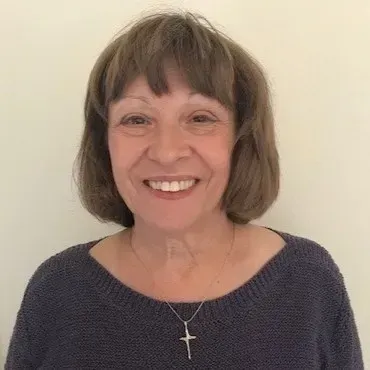 Smiling woman with short brown hair wearing a dark sweater and cross necklace against a plain wall