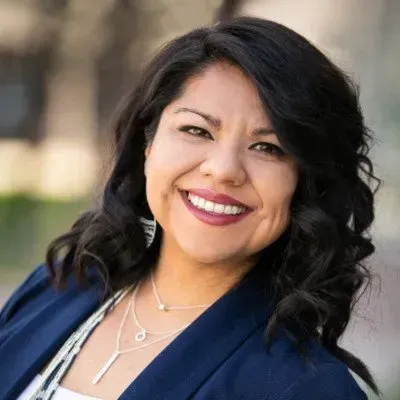 Smiling woman in a blue blazer with curly dark hair, standing outdoors.
