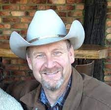 Smiling man in a white cowboy hat and brown jacket sitting on a porch with a brick wall background