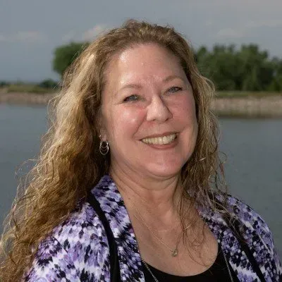 Smiling woman with wavy hair in a purple patterned top, standing by the water outdoors