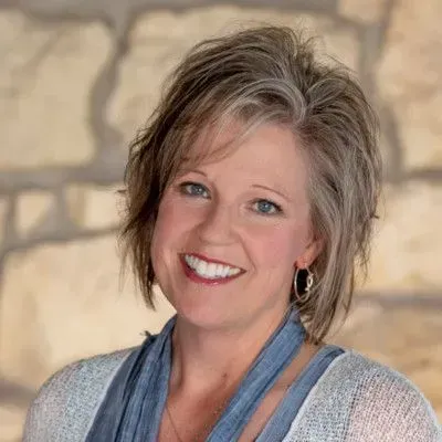 Smiling woman with short highlighted hair in a blue top and light cardigan against a stone wall
