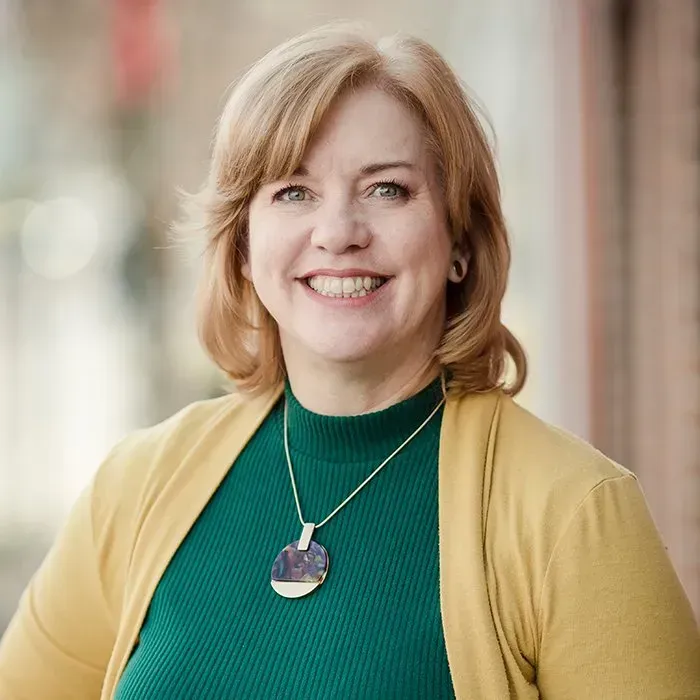 Smiling woman in a teal top and mustard cardigan, wearing a pendant necklace, standing outdoors