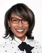 Smiling person with dark hair and glasses, wearing a white polka-dot blouse with a black bow.